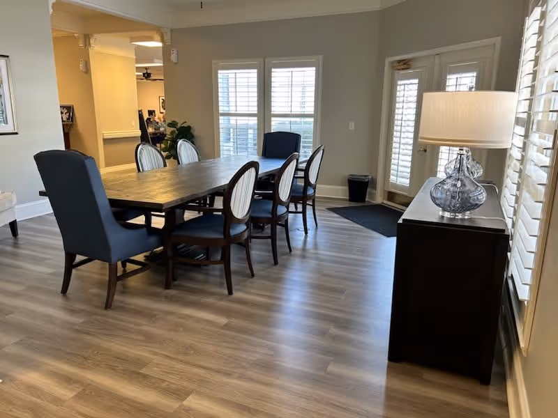 A dining area in a senior living facility with a long wooden table surrounded by six chairs, including two blue upholstered chairs at each end. The room has light wood flooring, large windows with white blinds, and a glass door leading outside. A dark wooden sideboard with a decorative glass lamp is positioned against the wall on the right.