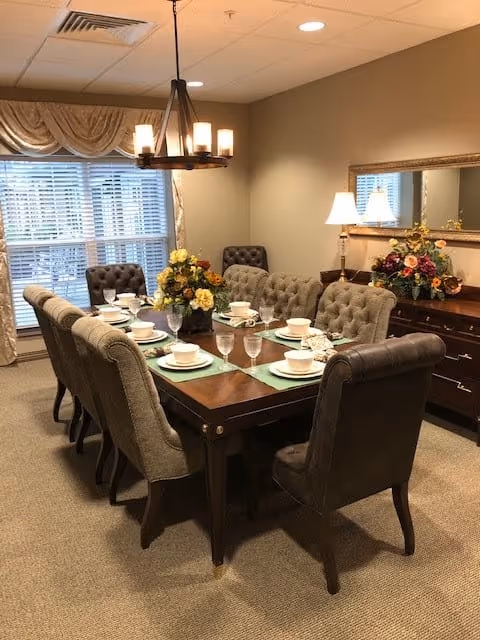A formal dining room with a long wooden table set for eight people. The table has place settings with white bowls, plates, and glasses on green placemats. There are upholstered chairs around the table. A chandelier with six lights hangs above the table. A large window with blinds and draped curtains is in the background. A sideboard with a lamp and floral arrangements is against the wall, beneath a large mirror.