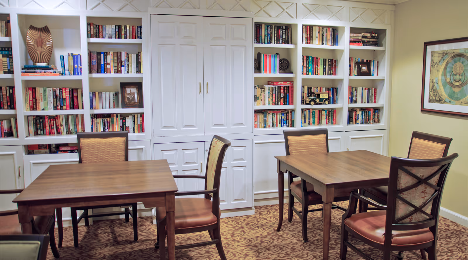 A cozy room with two wooden tables and several cushioned chairs arranged around them. The back wall features built-in white bookshelves filled with books and decorative items. A framed artwork hangs on the right wall, and the floor is carpeted with a patterned design.