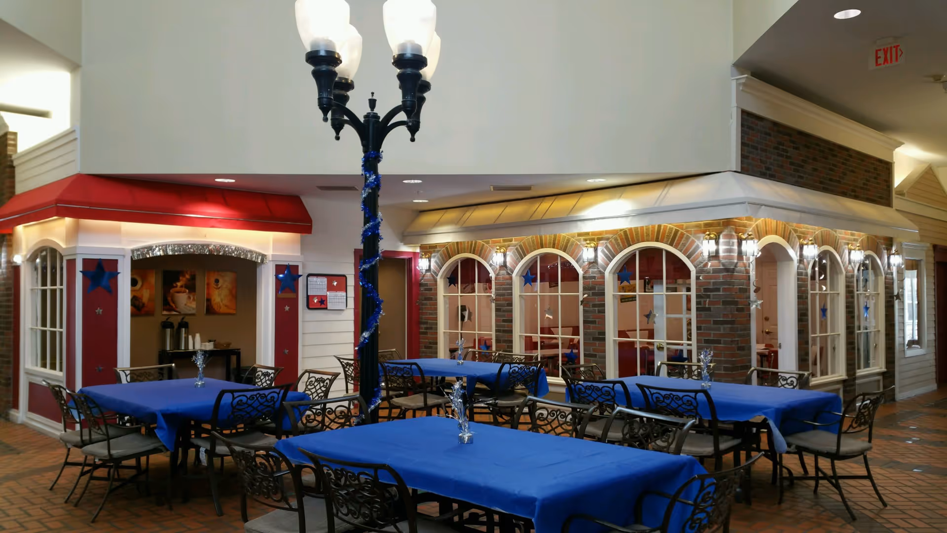 Indoor dining area with several tables covered in blue tablecloths and black wrought iron chairs. The space features decorative storefront-style walls with windows and red and beige awnings. A tall black lamp post with three lights stands in the center, wrapped with blue garland. The floor is tiled with a brick pattern, and there is an exit sign visible in the background.