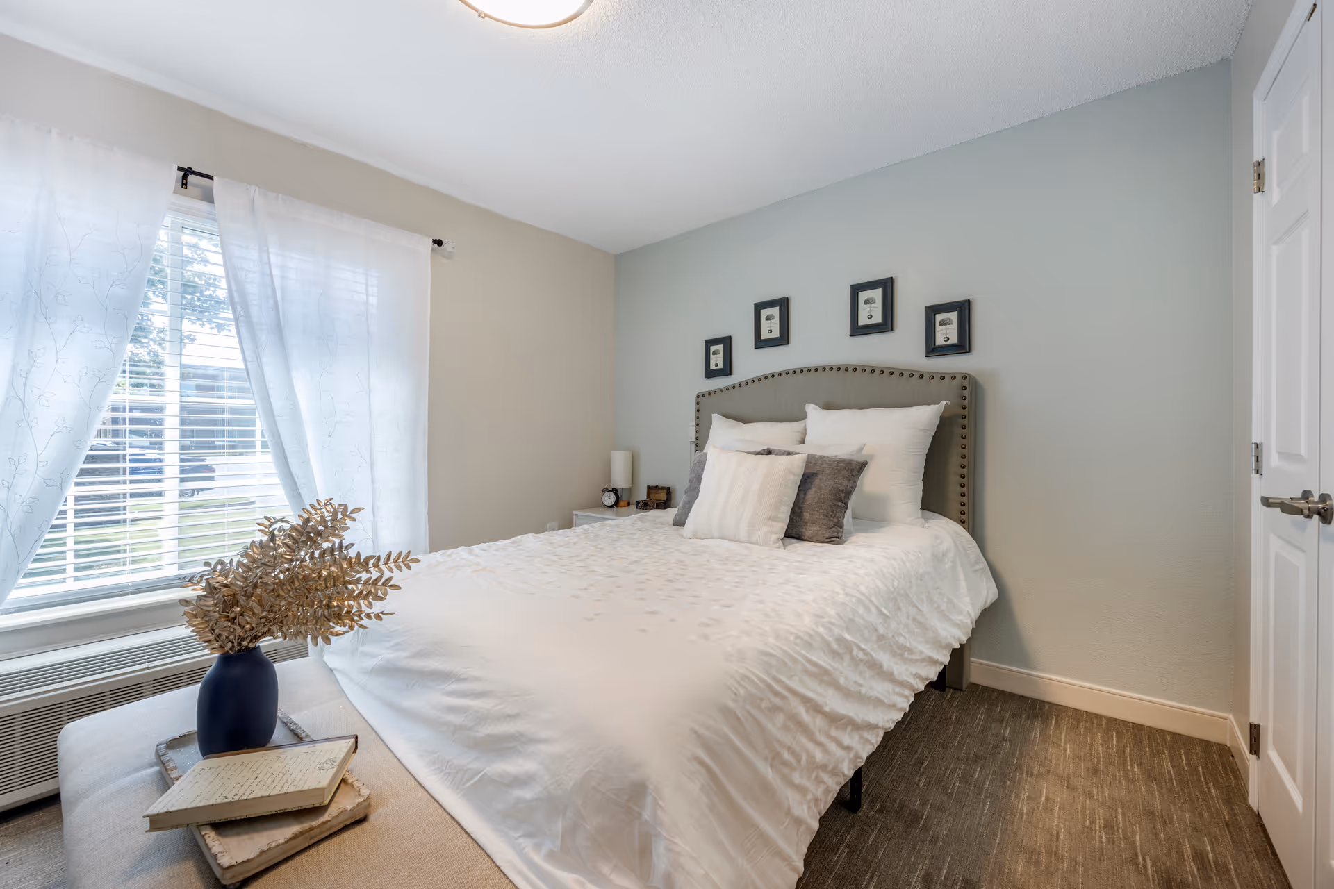 A neatly made bed with white linens and decorative pillows in a bright bedroom with a window, curtains, and a bench holding a vase and books.