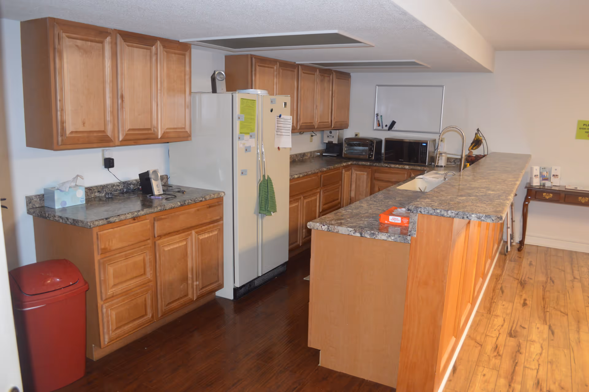 Interior view of a kitchen area with wooden cabinets, a white refrigerator, a countertop with a sink, a microwave, a toaster oven, and a red trash bin. The floor is wooden, and there is a small table with brochures and a sign on the wall in the background.
