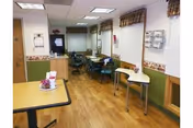 Indoor dining area in a care facility with tables and chairs, a serving counter, and wood-look flooring.