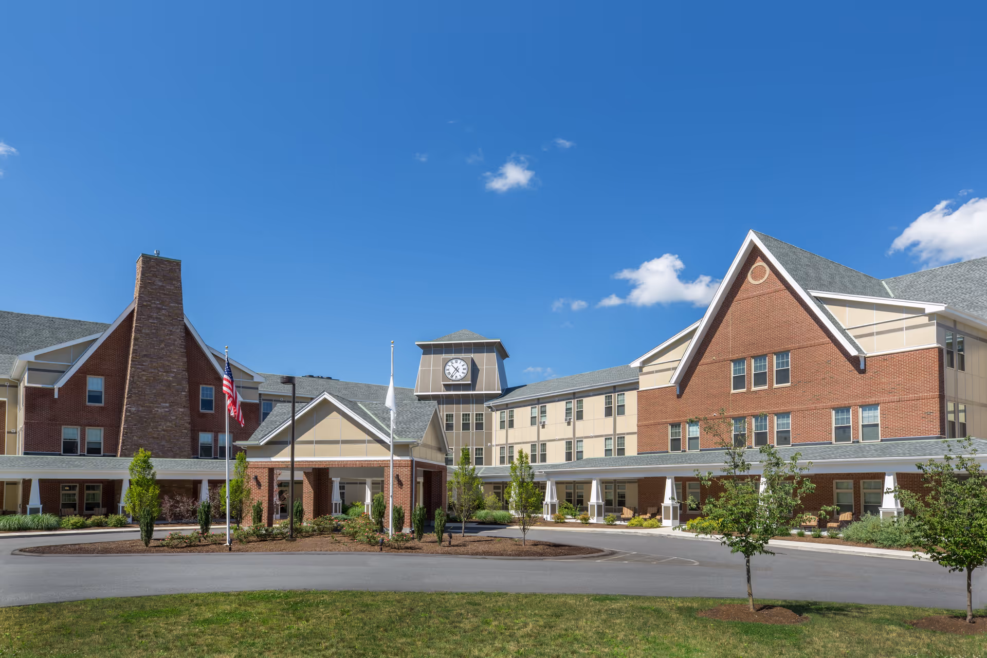 Exterior view of a large senior living facility building with a clock tower, brick and beige siding, a covered entrance, and landscaped grounds under a clear blue sky.