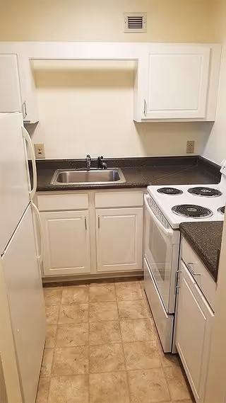 Small kitchen area with white cabinets, a white refrigerator on the left, a stainless steel sink in the center under a vent, and a white electric stove with coil burners on the right. The countertop is dark and the floor has beige tiles.
