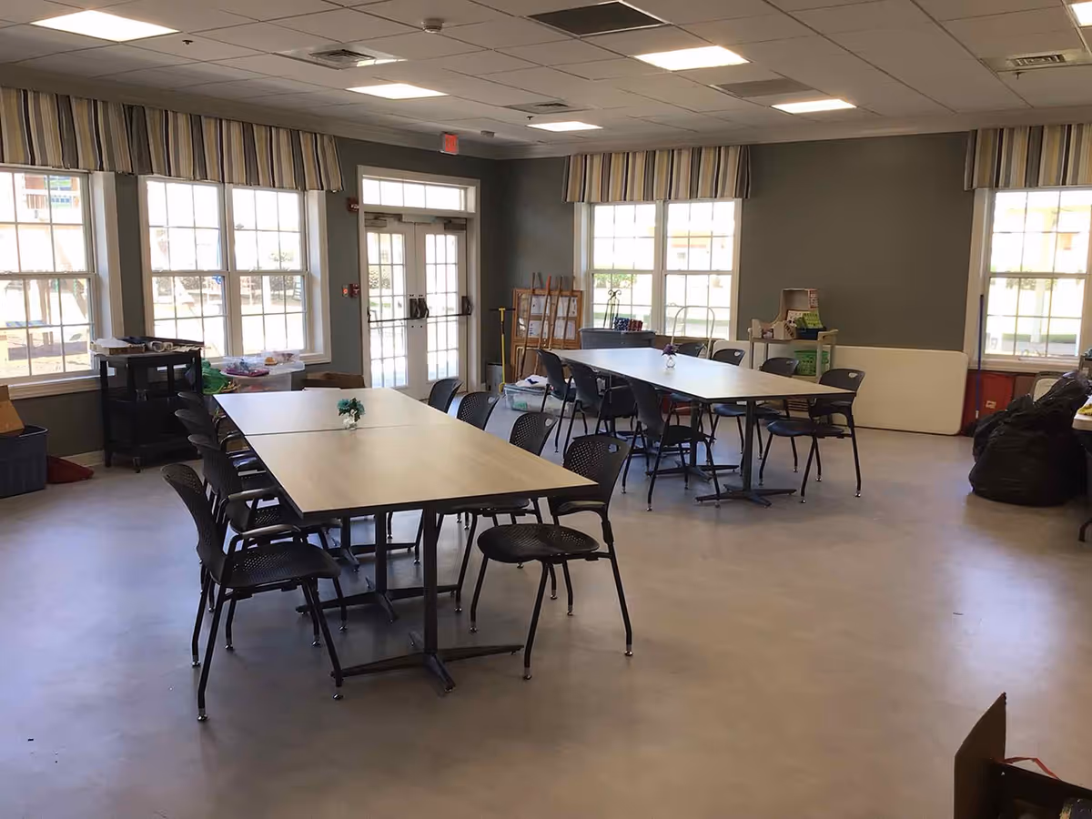 Bright community activity/dining room with multiple rectangular tables and chairs and large windows.