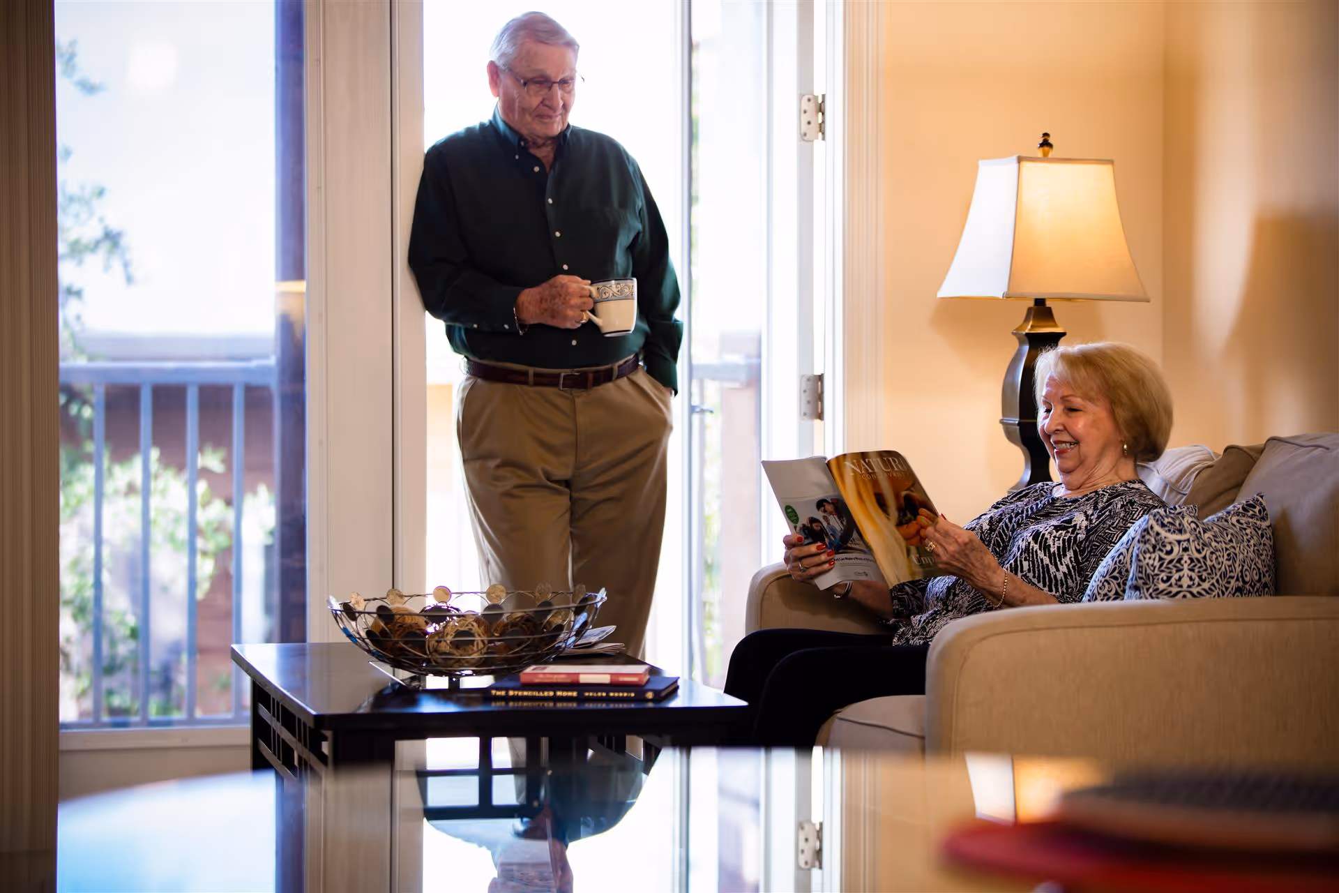 An elderly man holding a coffee mug stands near a glass door, looking at an elderly woman sitting on a beige couch reading a magazine. The room is warmly lit with a table lamp and has a coffee table with decorative items in front of the couch.