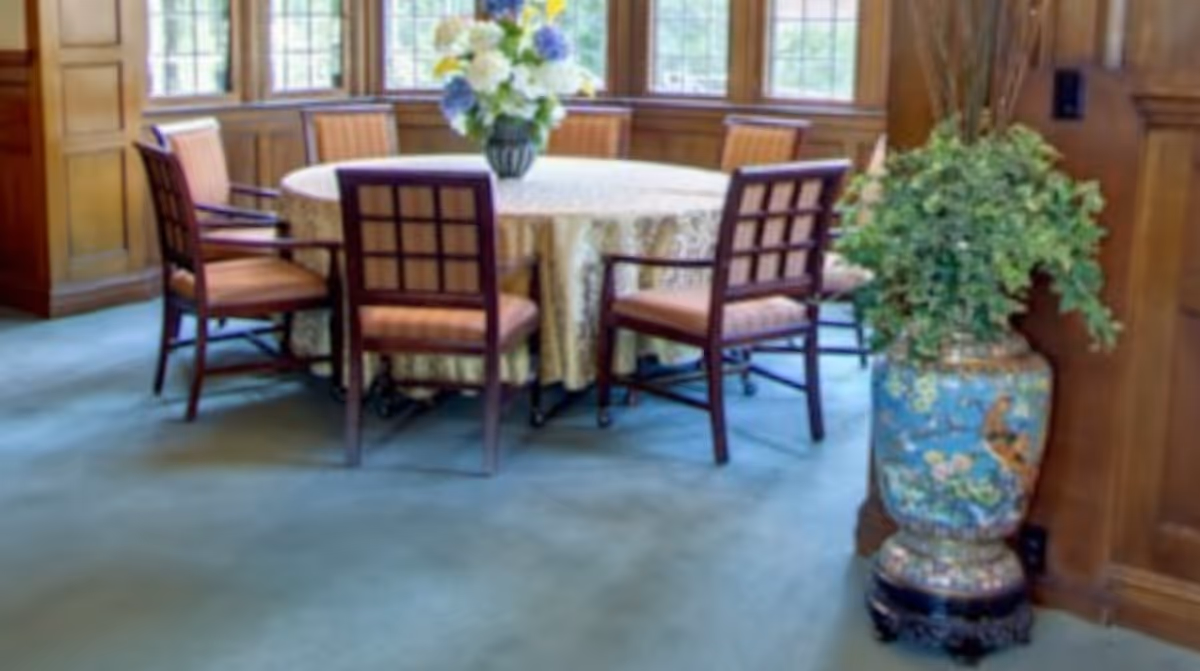 Round table with a tablecloth and vase of flowers surrounded by wooden chairs in a sunlit room with wood-paneled walls and a decorative potted plant.