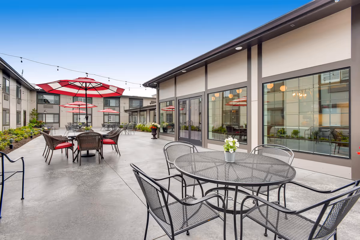 Outdoor patio area at Cascade Inn Assisted Living Community with multiple metal tables and chairs. Some tables have red and white striped umbrellas. The patio is surrounded by the building with large windows reflecting the outdoor furniture and umbrellas. String lights are hung above the seating area.
