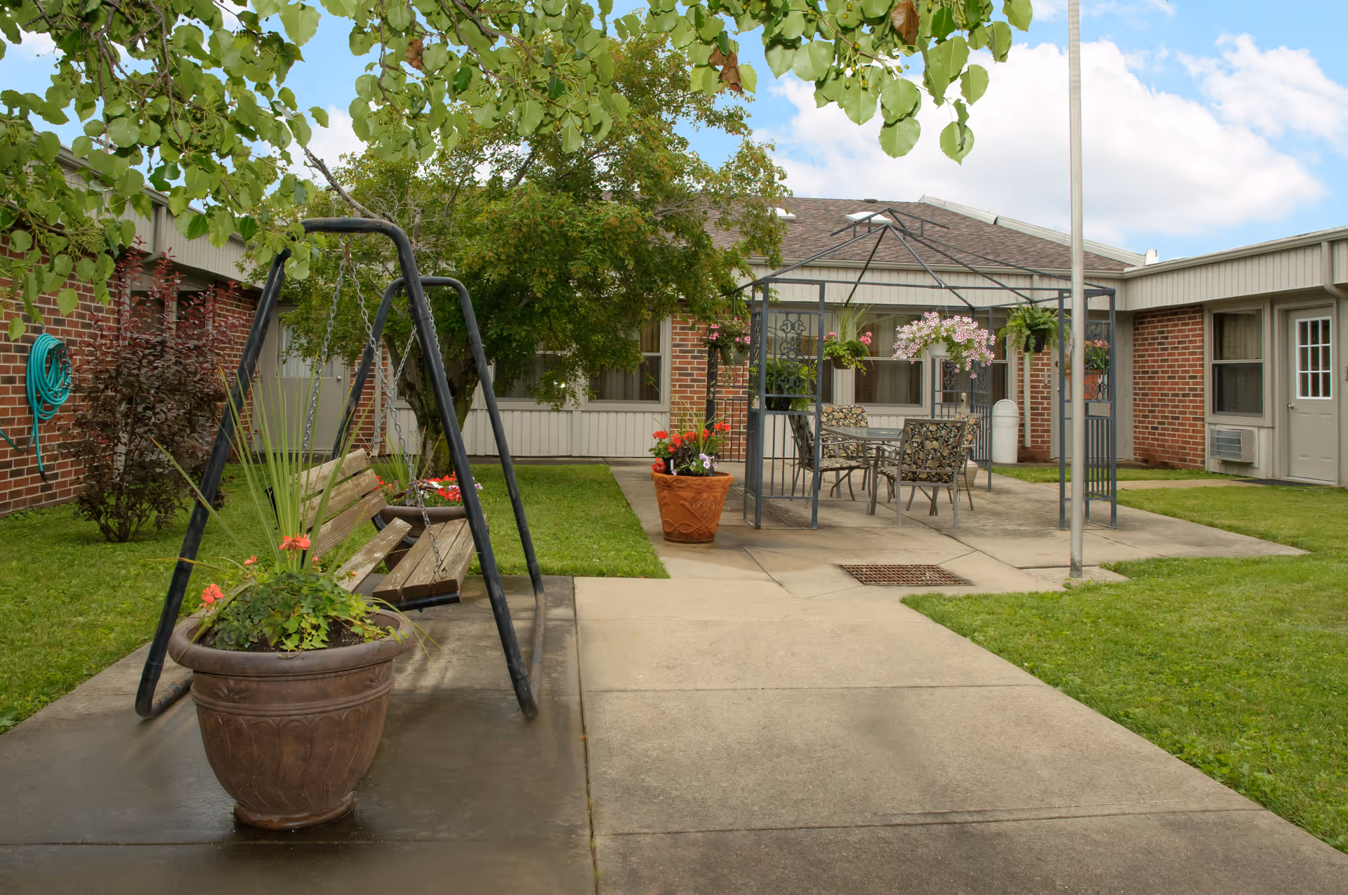 Outdoor courtyard area with a swing bench, potted plants, a metal gazebo with a table and chairs underneath, surrounded by a brick building with windows and doors, green grass, and a tree providing shade.