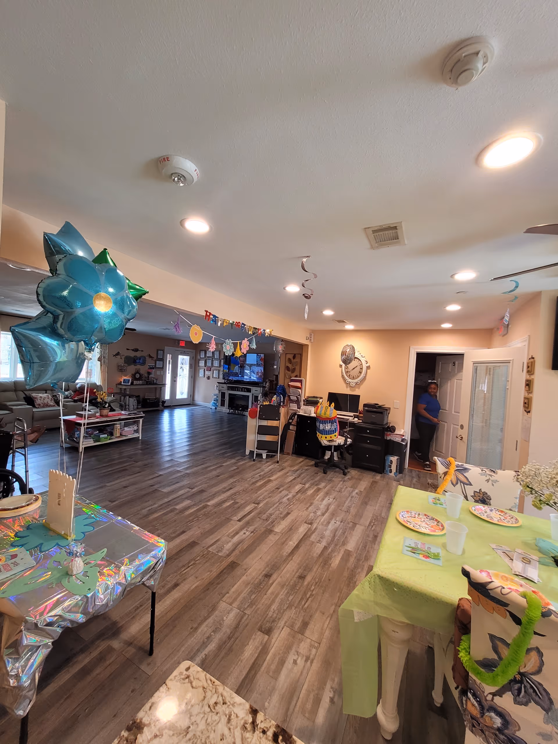 Interior view of a senior living facility common area decorated for a celebration with balloons, colorful banners, and tables set with plates and cups. The room has wood flooring, recessed ceiling lights, and a seating area with couches and a television in the background. A person is standing near a doorway on the right side.