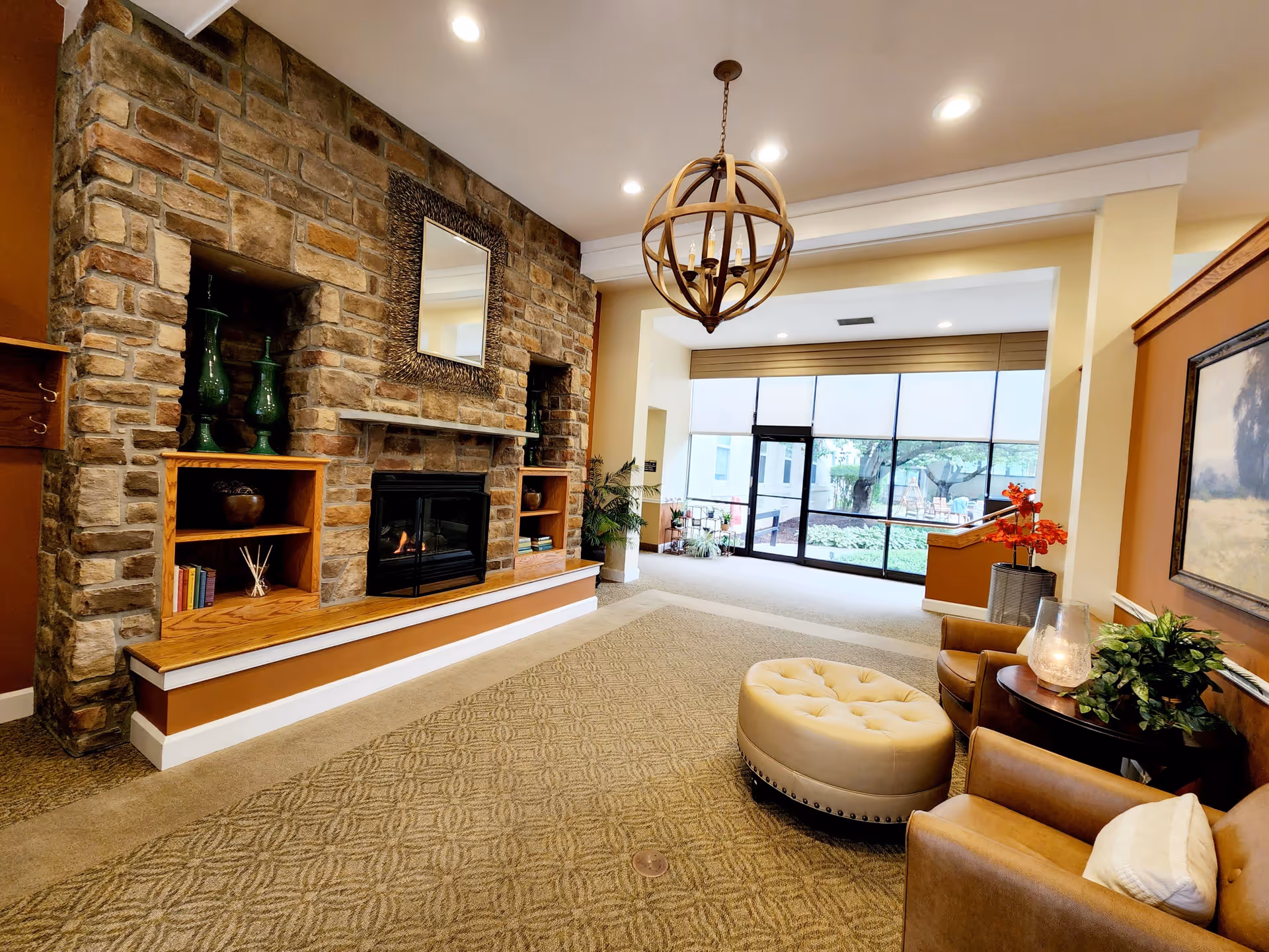 A cozy senior living facility common area featuring a stone fireplace with built-in wooden shelves holding decorative items and books. The room has a patterned carpet, two brown leather armchairs with a round beige ottoman in between, a small table with a candle and plant, and a large window with a view of an outdoor garden area. A spherical chandelier hangs from the ceiling.