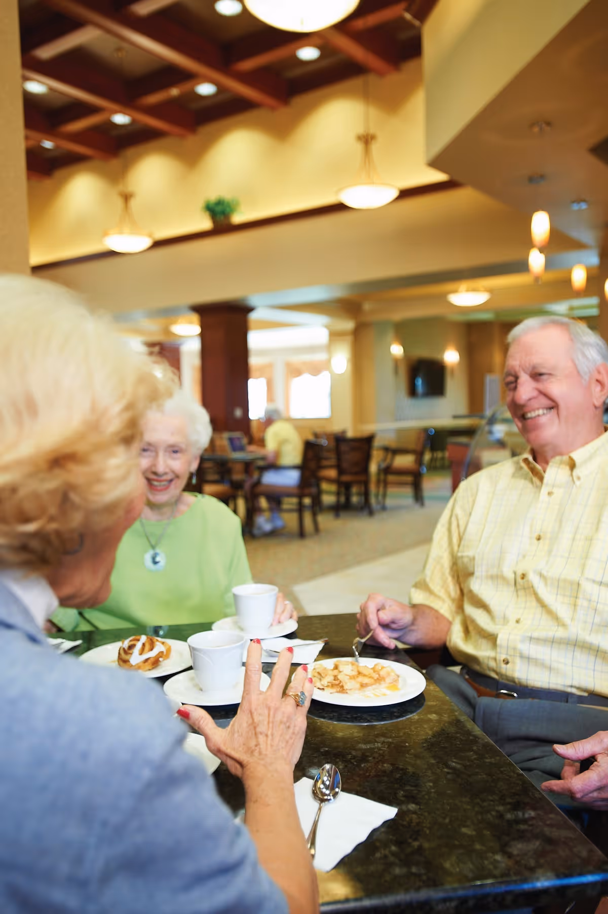 Three elderly people sitting around a table in a well-lit dining area, enjoying coffee and pastries while smiling and conversing.