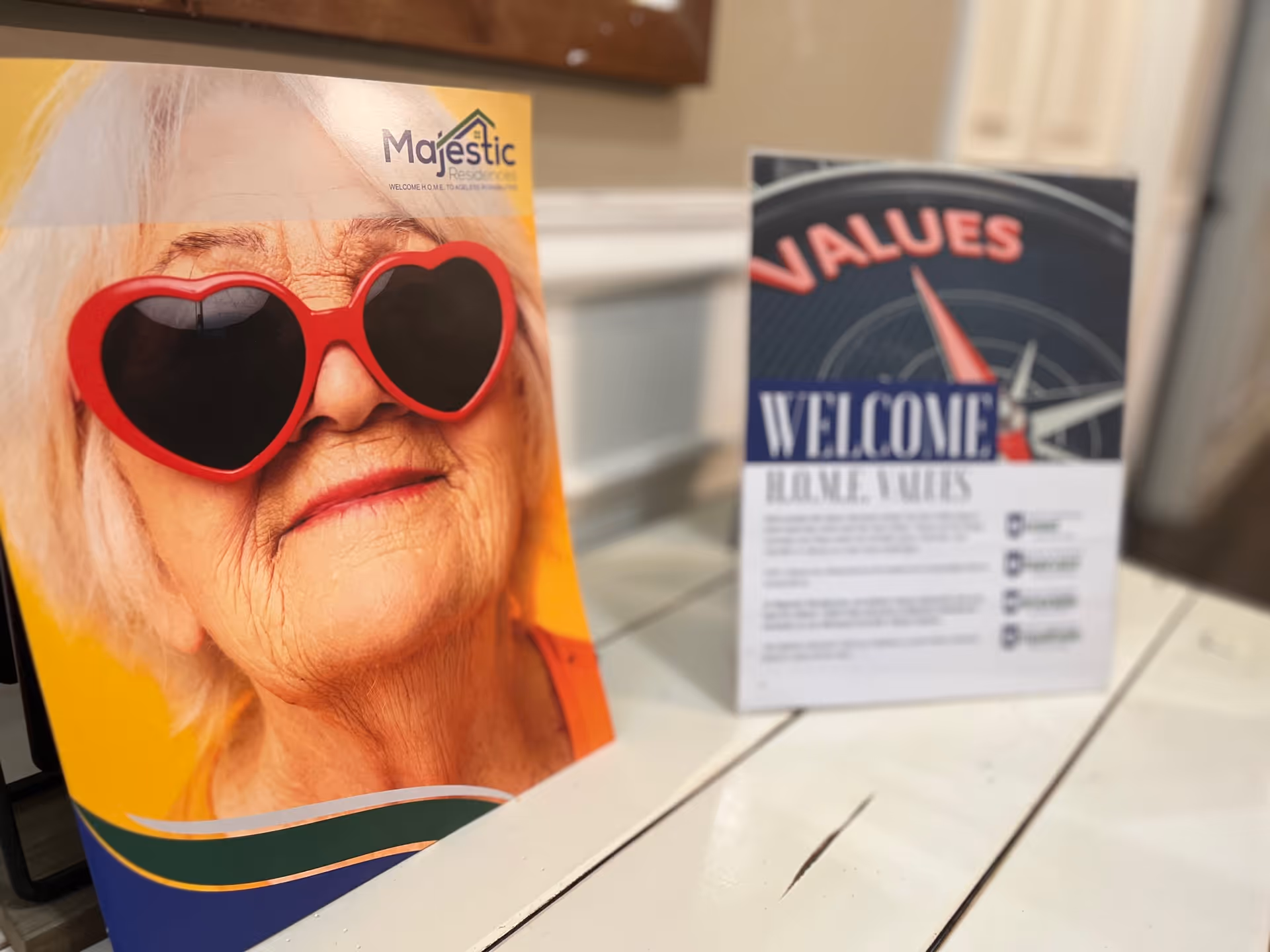 Two tabletop signs on a white wooden table, one showing a close-up of an elderly woman wearing red heart-shaped sunglasses and the other a 'Welcome' values sign.