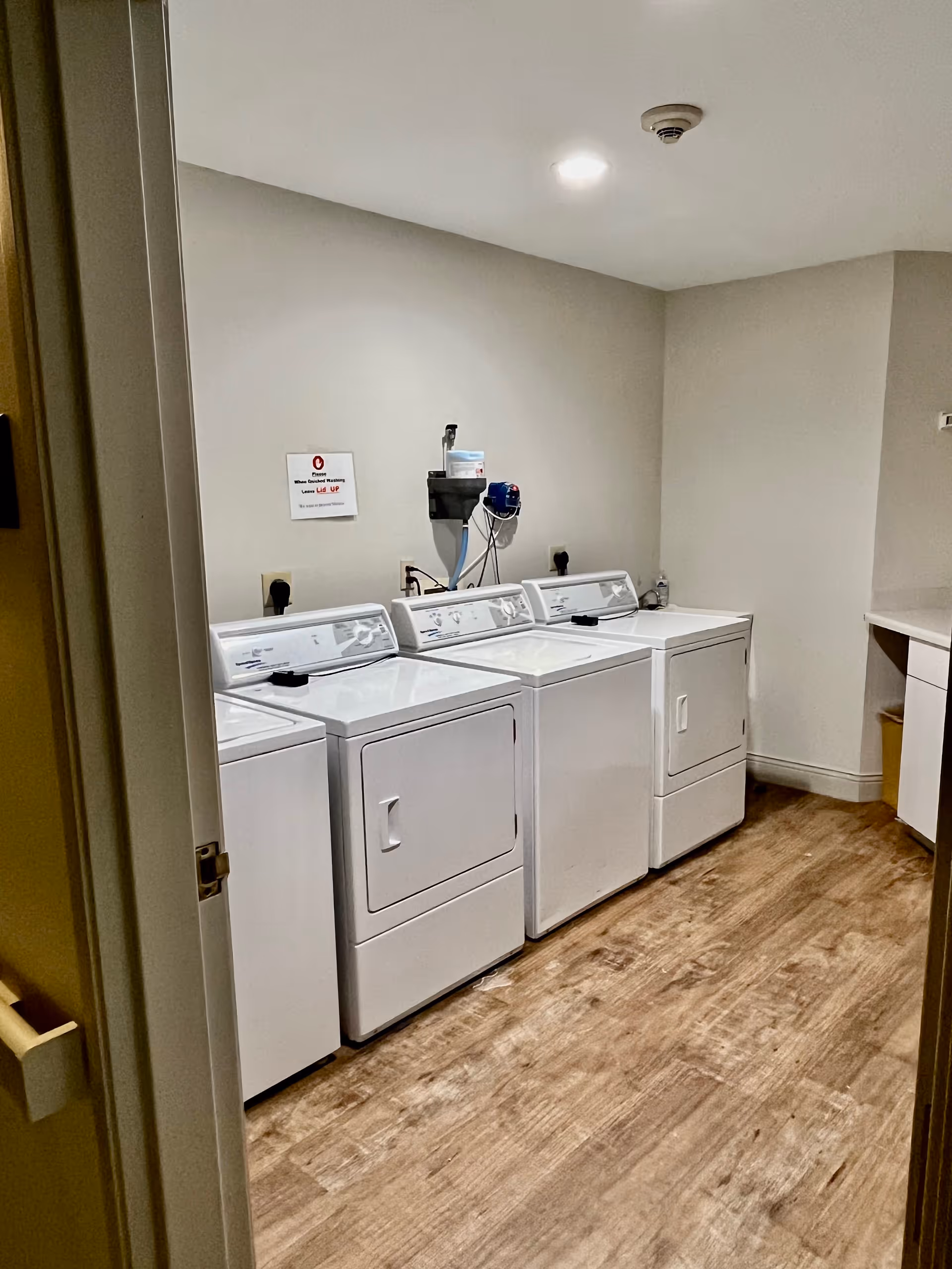 Laundry room with three white washing machines and dryers lined up against a beige wall with a wooden floor. There is a small sign on the wall above the machines and a countertop with cabinets on the right side.