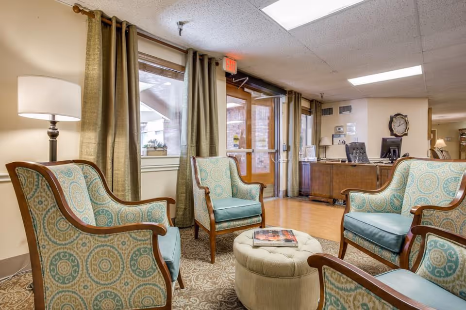 A cozy seating area in a senior living facility with four patterned armchairs arranged around a round tufted ottoman with magazines on top. The room has beige walls, green curtains, a floor lamp, and a wooden desk with a computer in the background near a glass door entrance.