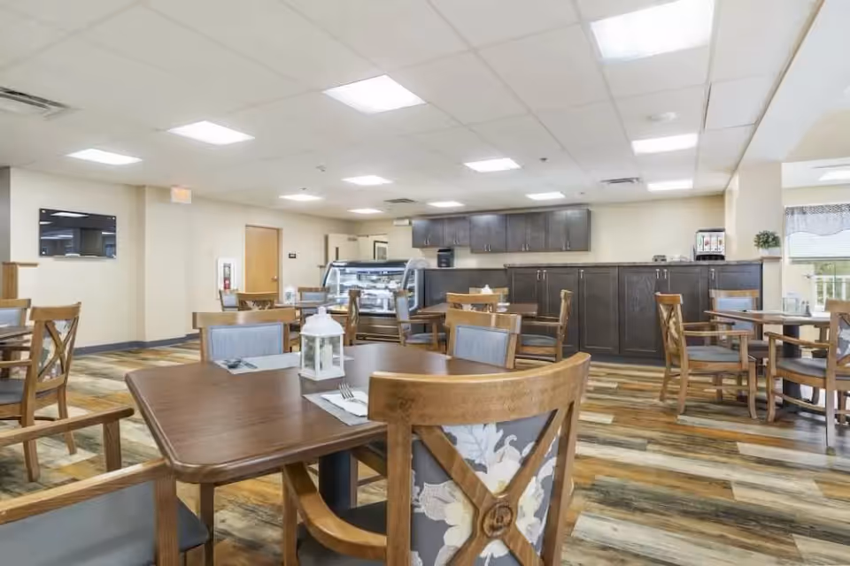 A bright and spacious dining room with wooden tables and chairs arranged neatly. The room features a display case with food items, dark wooden cabinets along the back wall, and a window with a valance letting in natural light. The floor has a wood-like pattern and the ceiling has recessed lighting panels.