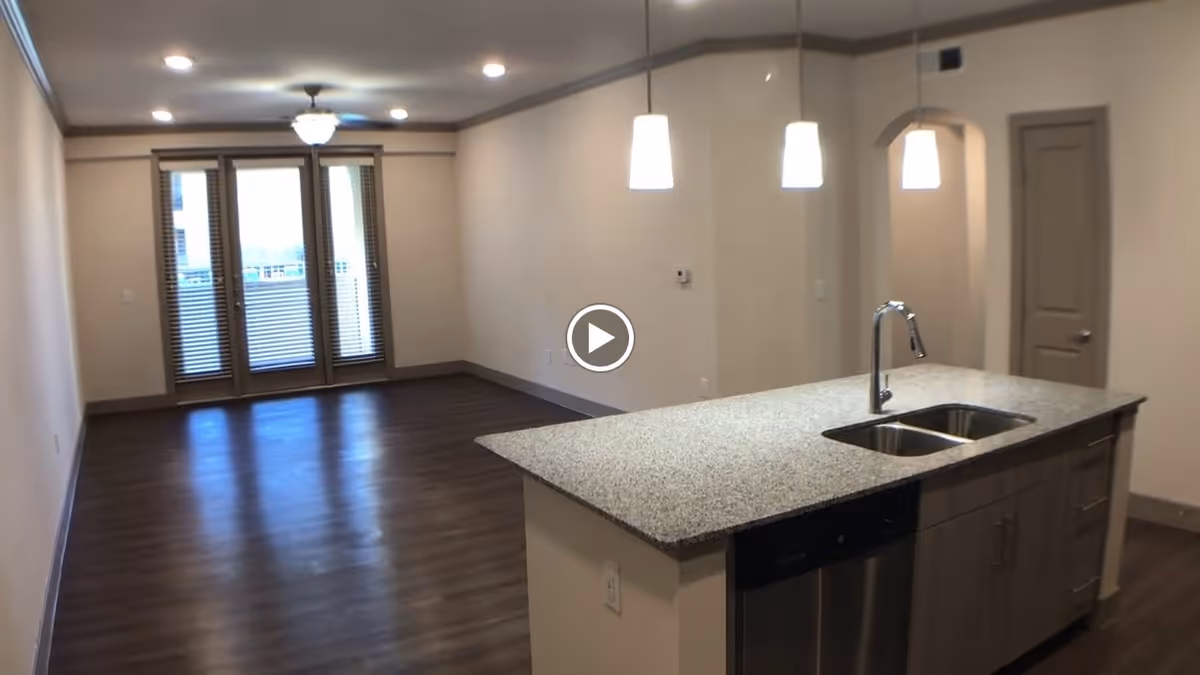 Open-plan interior showing a kitchen island with sink and pendant lights opening into a living area with hardwood floors and French doors with blinds.