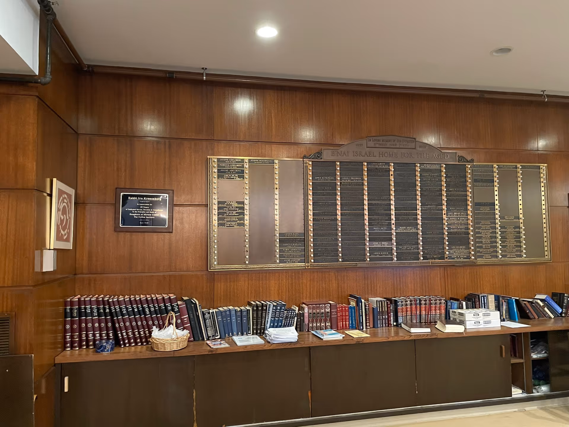 Interior view of a room with wood-paneled walls featuring a large commemorative plaque and a shelf below filled with numerous books and some papers. The plaque is titled 'Bnai Israel Home for the Aged'. There is also a smaller plaque on the left wall and a framed artwork above a light switch.