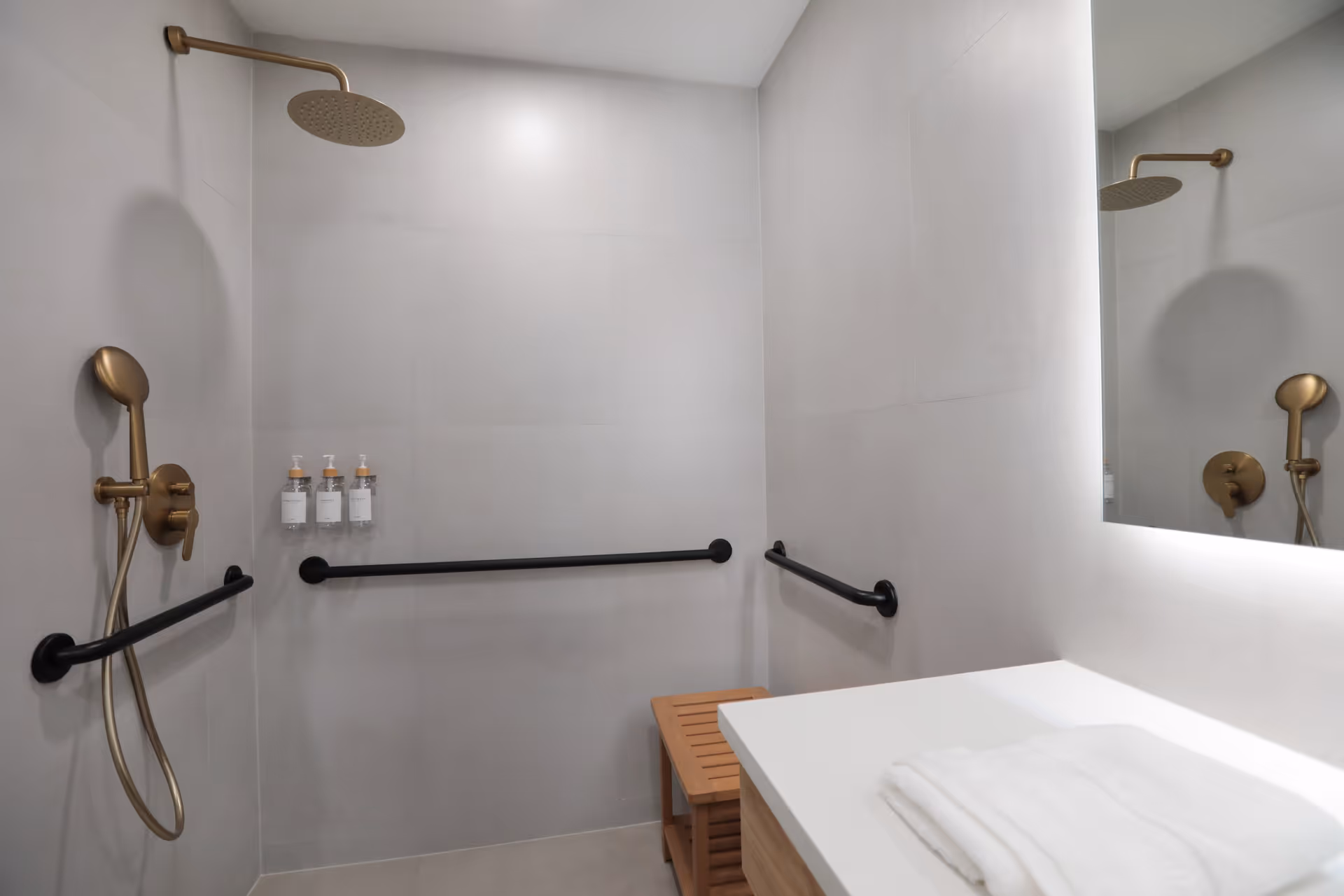 Modern accessible shower area with gold shower fixtures, black grab bars on the walls, a wooden bench, and a white countertop with folded towels. The walls are tiled in light gray, and a large mirror is visible on the right side.