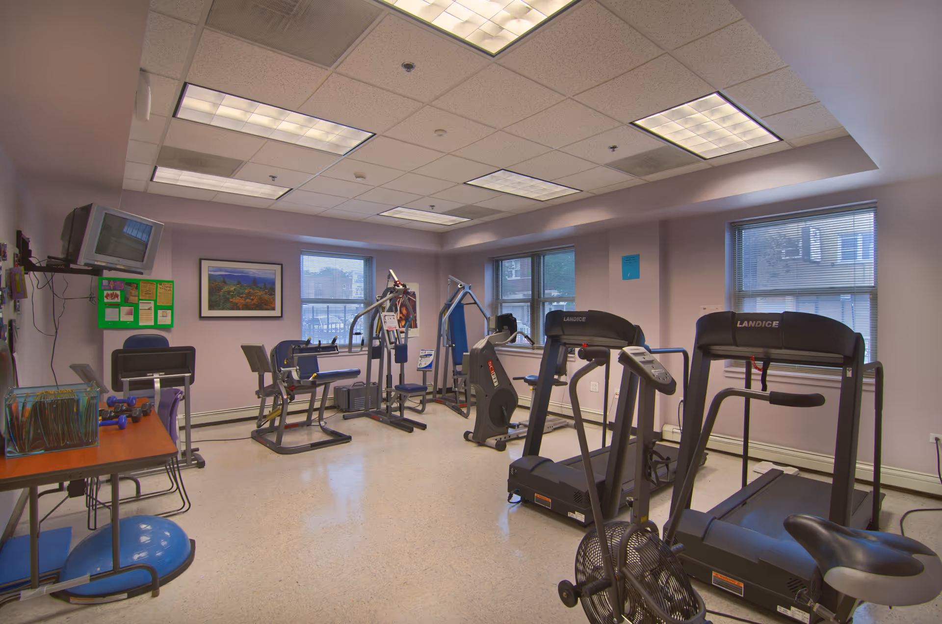 A fitness room in a senior living facility with exercise equipment including treadmills, stationary bikes, and strength training machines. The room has light purple walls, large windows with blinds, fluorescent ceiling lights, and a small desk with a TV mounted on the wall.