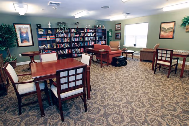 A spacious common room with patterned carpet, several wooden tables and chairs, a bookshelf filled with books, and comfortable armchairs near a large window. The walls are decorated with framed pictures and plants.