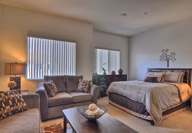 A cozy bedroom with a large bed covered in beige and brown bedding, a dark wooden dresser with decorative items, two windows with vertical blinds, a gray loveseat and armchair with patterned cushions, a coffee table with a decorative bowl, and a table lamp on a side table.