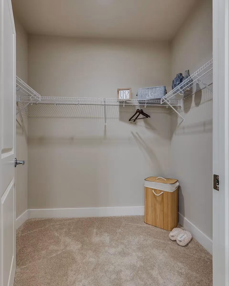 Empty walk-in closet with beige carpet, white wire shelving on three walls, a wooden hamper with a white cloth lining in the corner, two slippers on the floor, a small framed sign that says 'HOME', a gray fabric storage bin, and two decorative vases on the shelves.