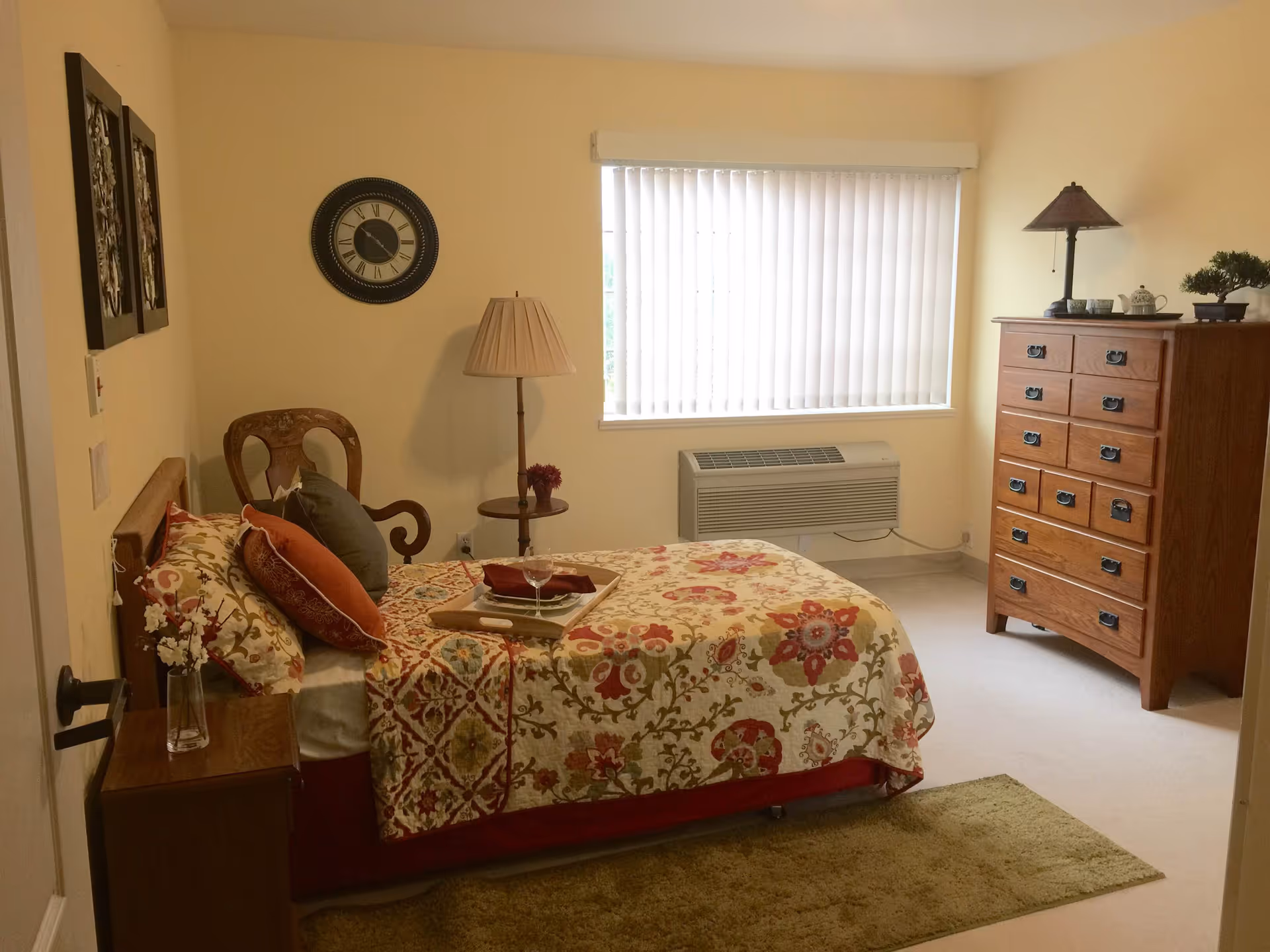 A neatly furnished bedroom with a floral bedspread, wooden dresser, chair, and a window with vertical blinds.