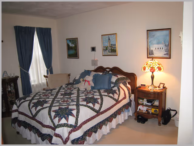 A cozy bedroom with a bed covered in a quilted bedspread featuring a star pattern. The bed has several pillows and a teddy bear on it. To the right of the bed is a wooden nightstand with a Tiffany-style lamp, a clock, and various small items. There are three framed pictures hanging on the wall above the bed. A window with blue curtains is on the left side of the room, and a chair is positioned near the window.