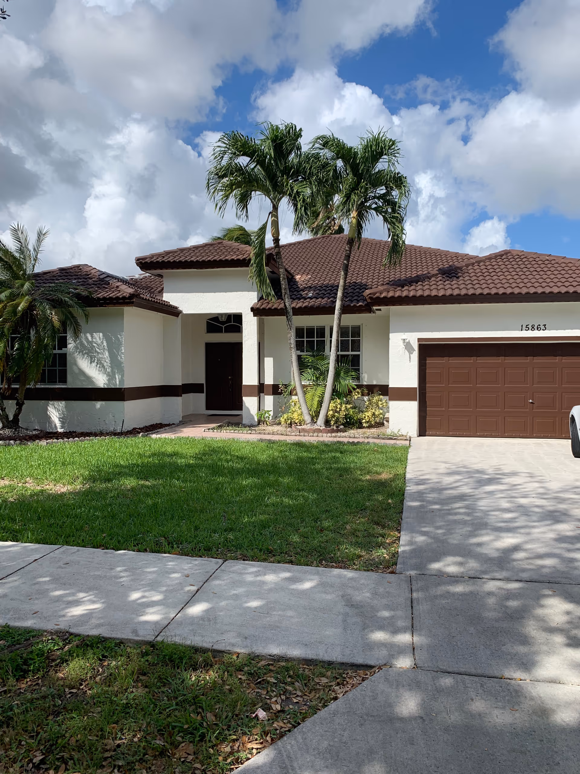 Front exterior view of a single-story house with a brown tiled roof, white walls with a brown horizontal stripe, a brown garage door, a front door with a small window above it, and two palm trees in the front yard. The sky is partly cloudy.