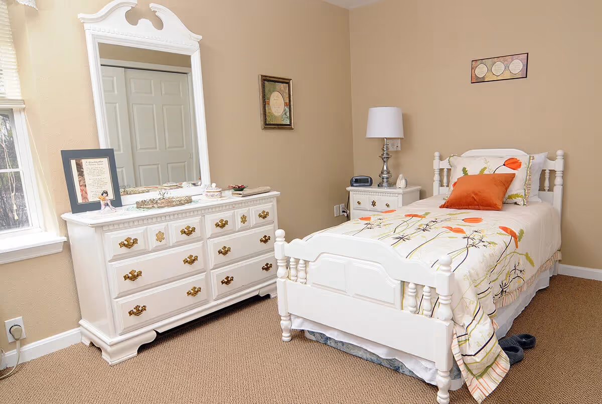 A cozy bedroom with a white wooden bed featuring floral bedding and an orange pillow. Next to the bed is a white nightstand with a silver lamp and a clock. Across from the bed is a white dresser with ornate gold handles and a large mirror. The walls are beige, and there are two framed pictures hanging. The room has a window with blinds partially open, letting in natural light.