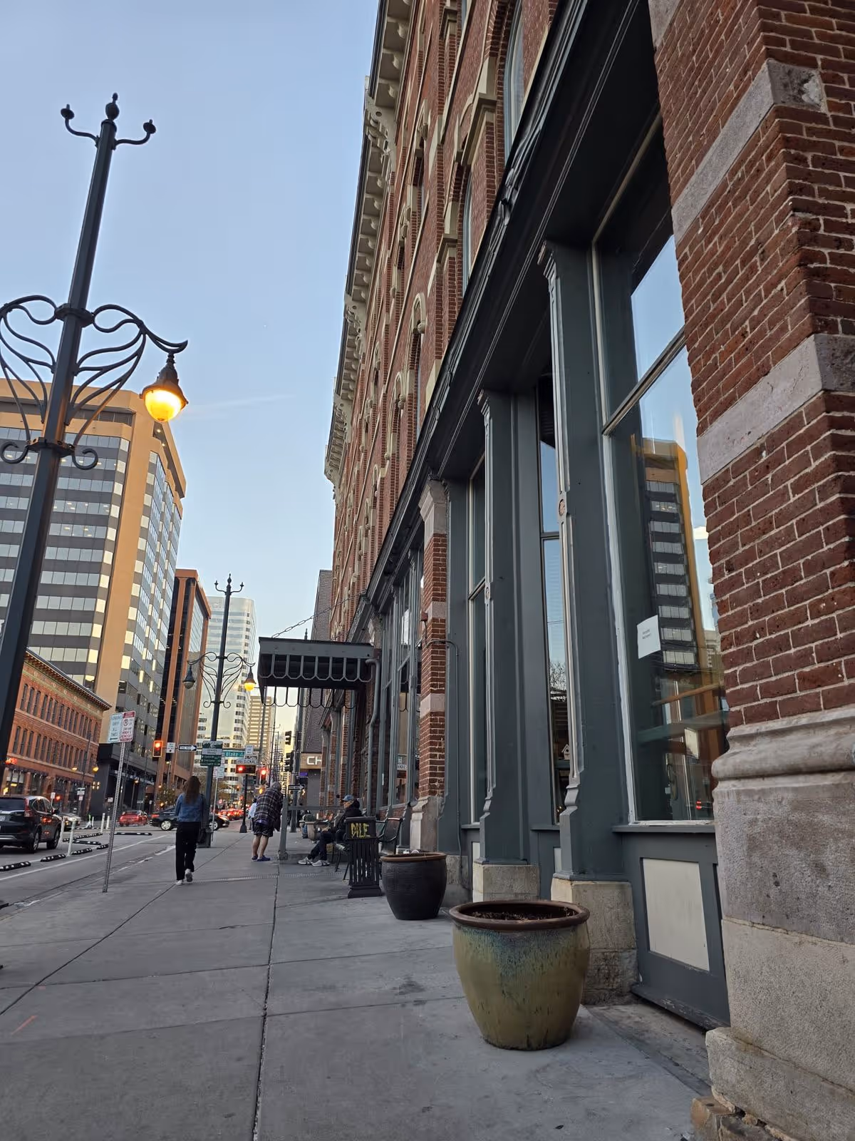 Sidewalk view of a multi-story brick building façade with storefront windows, planters, lampposts, and people walking on a city street.