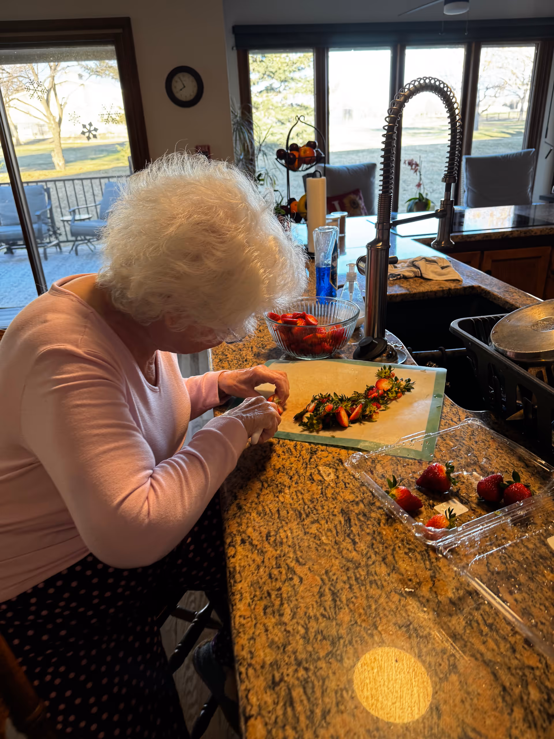 An elderly woman with white hair is sitting at a kitchen counter, preparing strawberries by removing their stems. The kitchen has a granite countertop with a cutting board, a bowl of strawberries, a sink with a modern faucet, and a drying rack with dishes. Large windows in the background show an outdoor patio and trees.