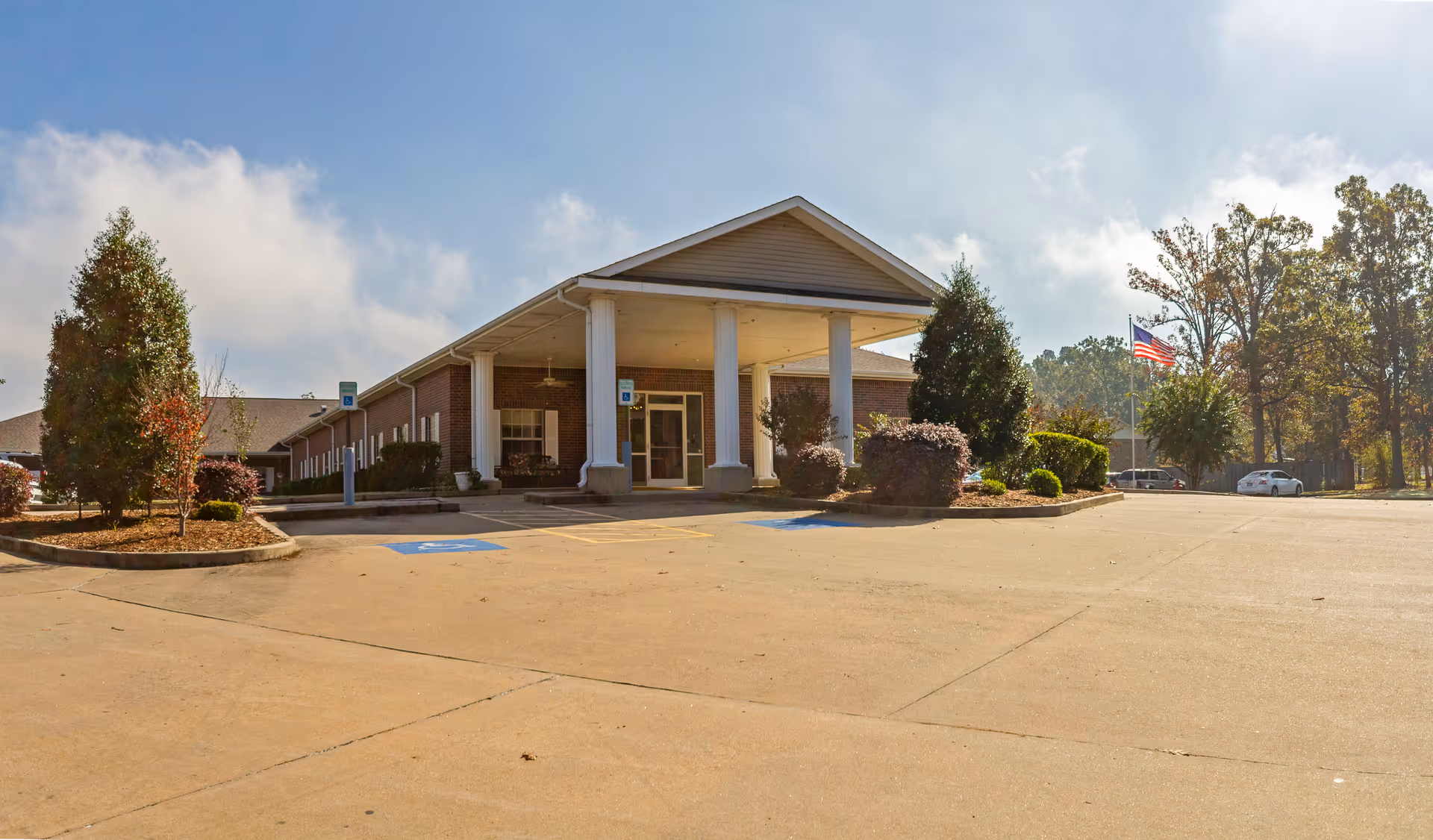 Exterior view of The Blossoms at White Hall Rehab & Nursing Center showing a single-story brick building with a covered entrance supported by white columns. The parking lot in front has designated handicap parking spaces, and there are landscaped areas with trees and shrubs around the building. An American flag is visible on a flagpole to the right.