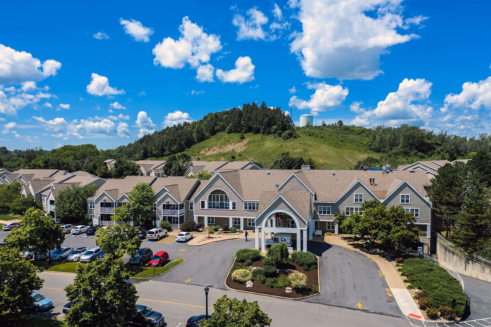 Exterior view of The Commons on St. Anthony, a multi-building senior living campus with a curved entrance and parking lot, set against a grassy hill under a blue sky.