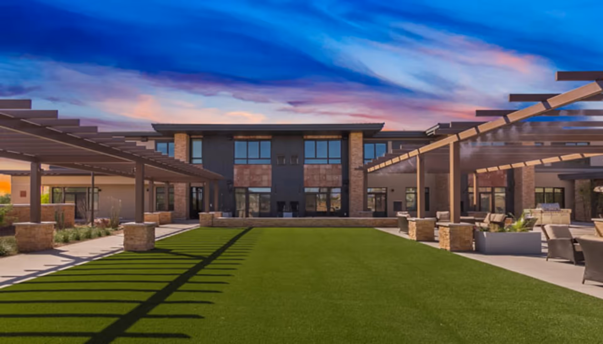 Outdoor courtyard area of a senior living facility with green artificial turf, shaded pergolas on both sides, stone pillars, seating areas with chairs and tables, and a modern two-story building in the background under a colorful sunset sky.