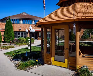 Outdoor courtyard at St. Clare Terrace featuring a wooden gazebo, paved walkway, and landscaped shrubs beside a brick building.