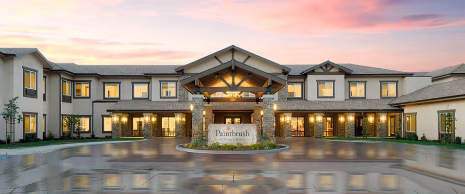 Front exterior view of Paintbrush Assisted Living facility at sunset with a circular driveway and illuminated entrance featuring stone pillars and a sign with the facility name.