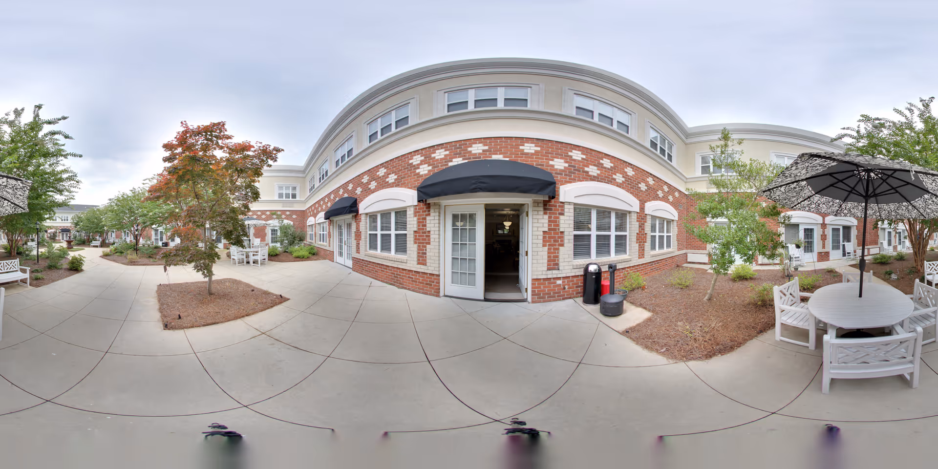 Outdoor courtyard area of a senior living facility with a curved brick and white building featuring multiple windows and black awnings. The courtyard has paved walkways, landscaped garden beds with small trees and shrubs, white outdoor tables and chairs, and umbrellas providing shade.