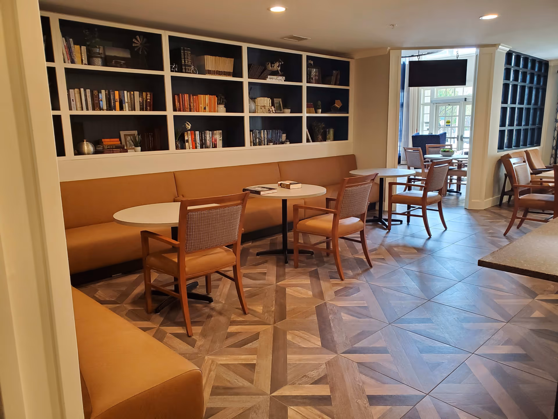 Cozy communal dining and reading area with round tables, wooden chairs, built-in bookshelves and banquette seating.