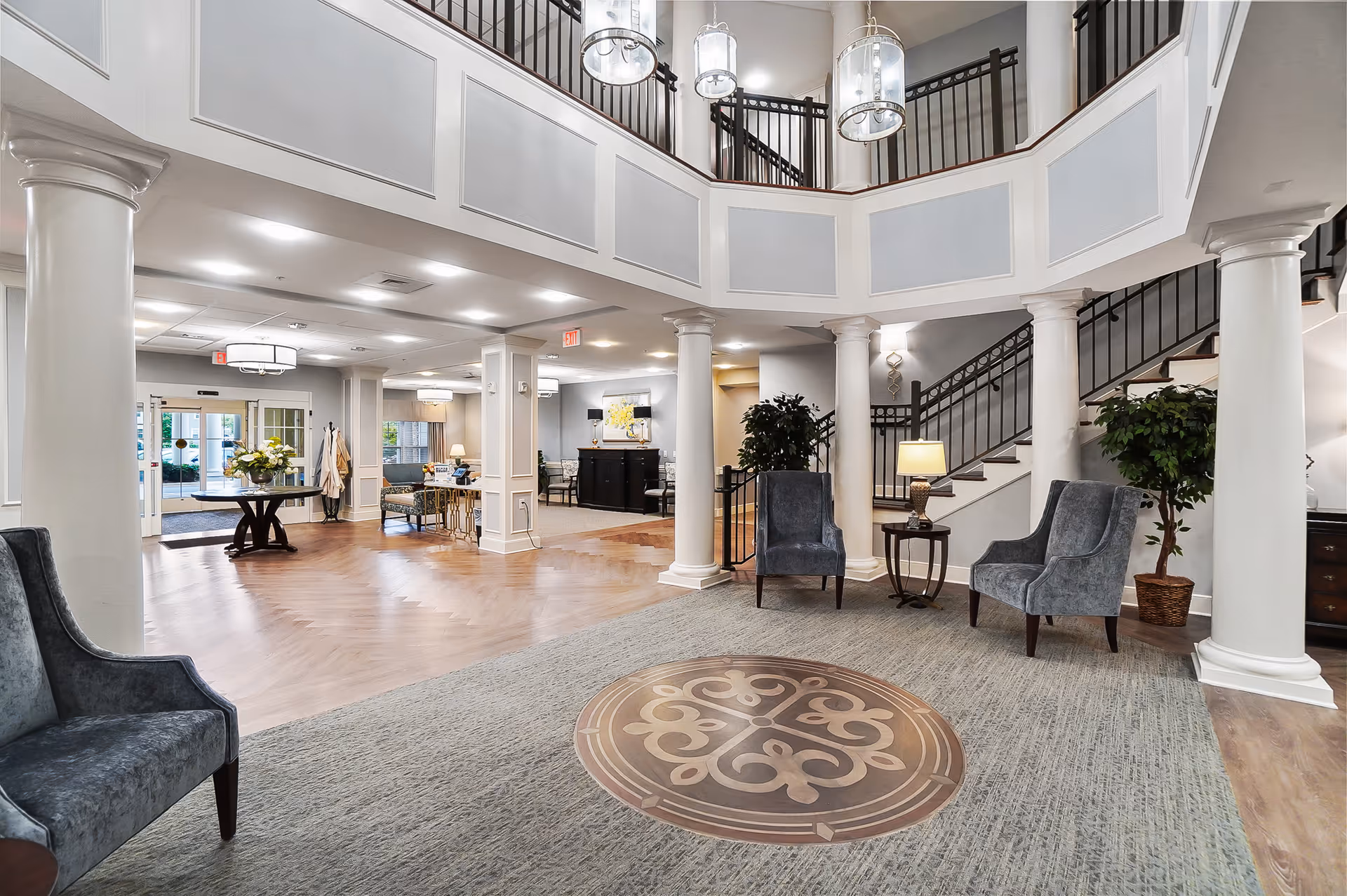 Spacious two-story lobby with seating, columns, a stairway and chandeliers in a senior living facility.