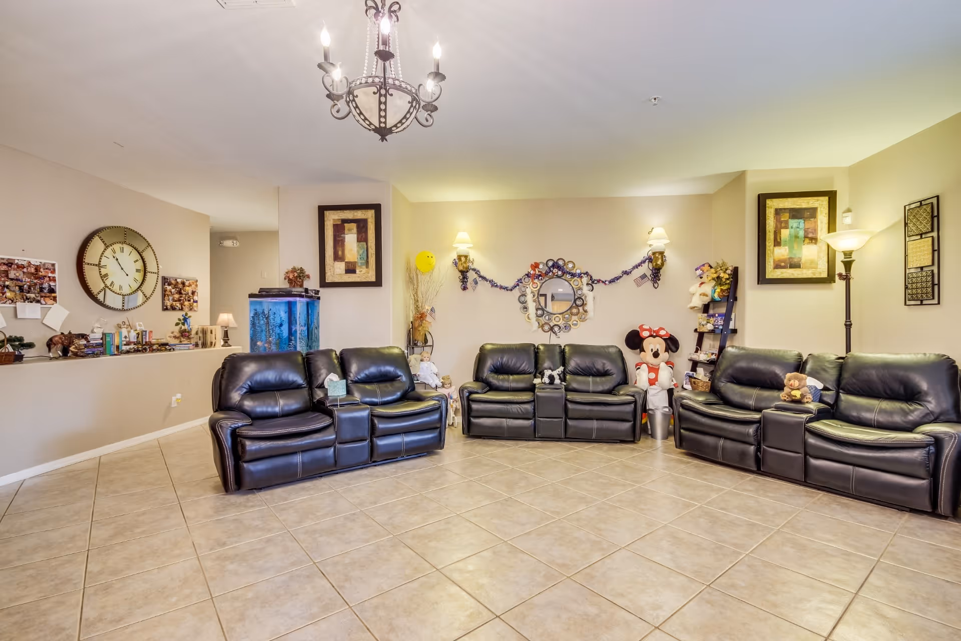 A spacious living room with three black leather reclining sofas arranged in a U-shape on a tiled floor. The walls are decorated with framed artwork, a large clock, a circular mirror with decorative plates around it, and various stuffed toys including a large Minnie Mouse doll. There is a chandelier hanging from the ceiling and a floor lamp in the corner. A fish tank is visible in the background along with a shelf holding more decorations and toys.