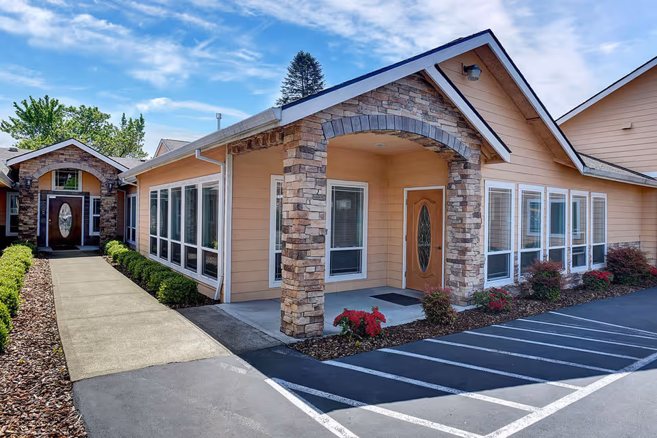 Exterior view of a residential care facility building with peach-colored siding and stone accents around the entrance. There is a concrete walkway leading to a wooden door with a decorative glass panel. The building is surrounded by small shrubs and flowers, and there is a parking area with marked spaces in front.