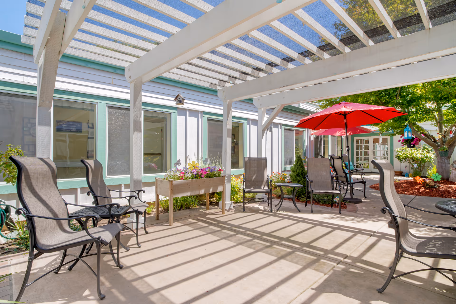 Outdoor patio area at Brookdale North Fremont with several metal and mesh chairs arranged under a white pergola. There is a planter box with flowers, a red umbrella providing shade over some chairs, and trees and shrubs surrounding the patio.