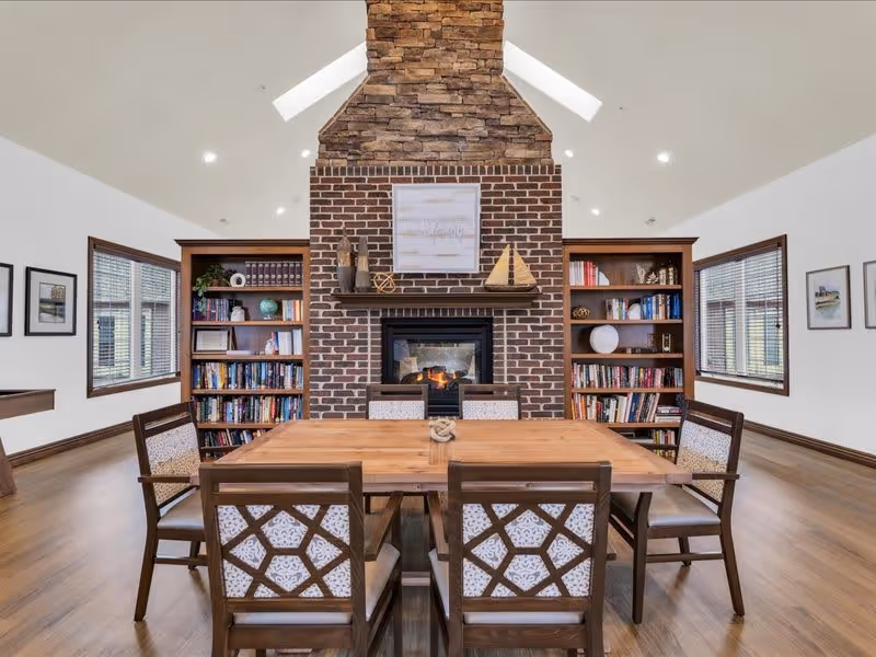 A cozy interior room with a wooden dining table surrounded by six chairs featuring patterned cushions. Behind the table is a brick fireplace with a stone chimney, flanked by two wooden bookshelves filled with books and decorative items. The room has large windows on both sides and a high ceiling with recessed lighting.