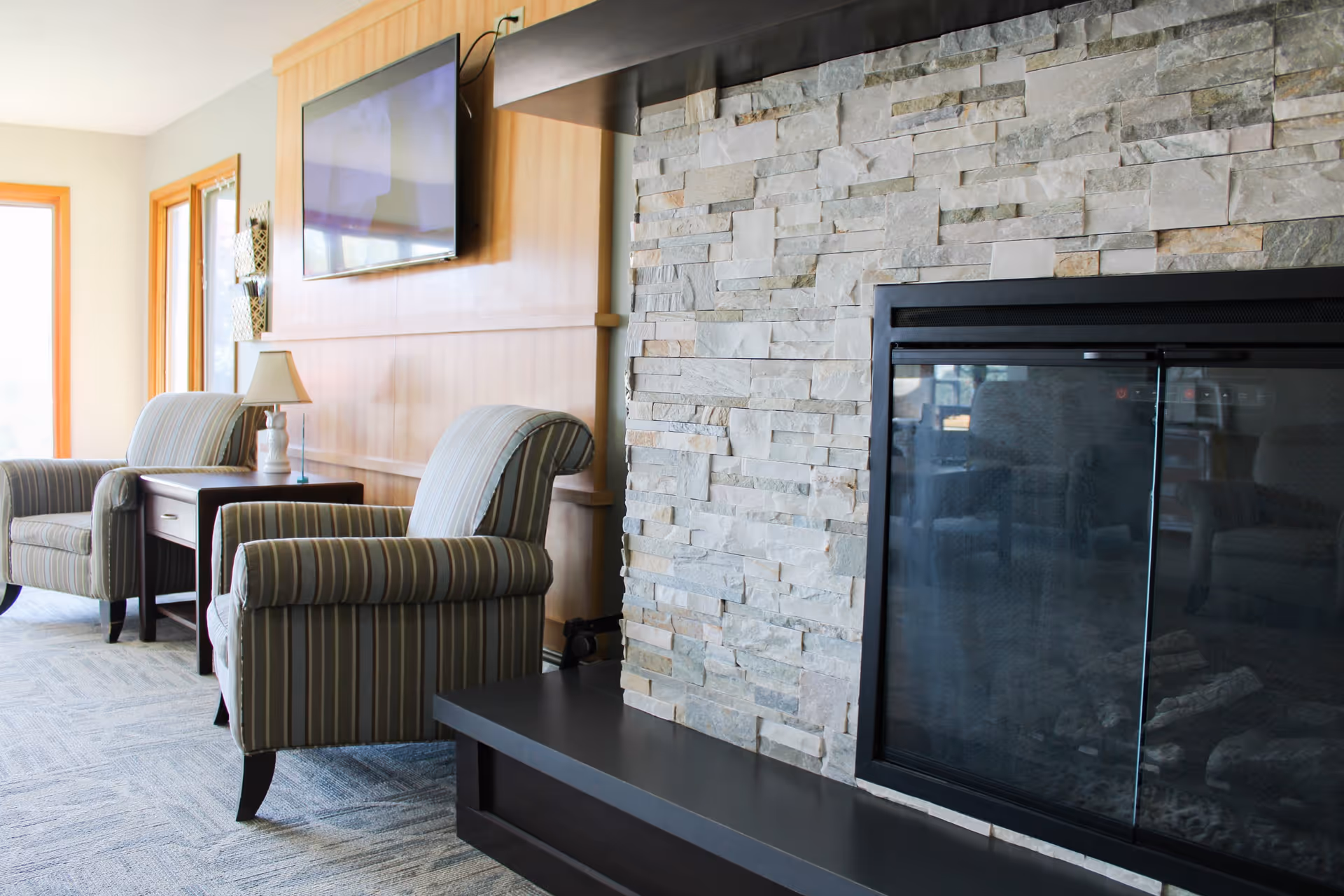 A cozy lounge area with two striped armchairs, a side table and lamp, a wall-mounted TV, and a stone fireplace.