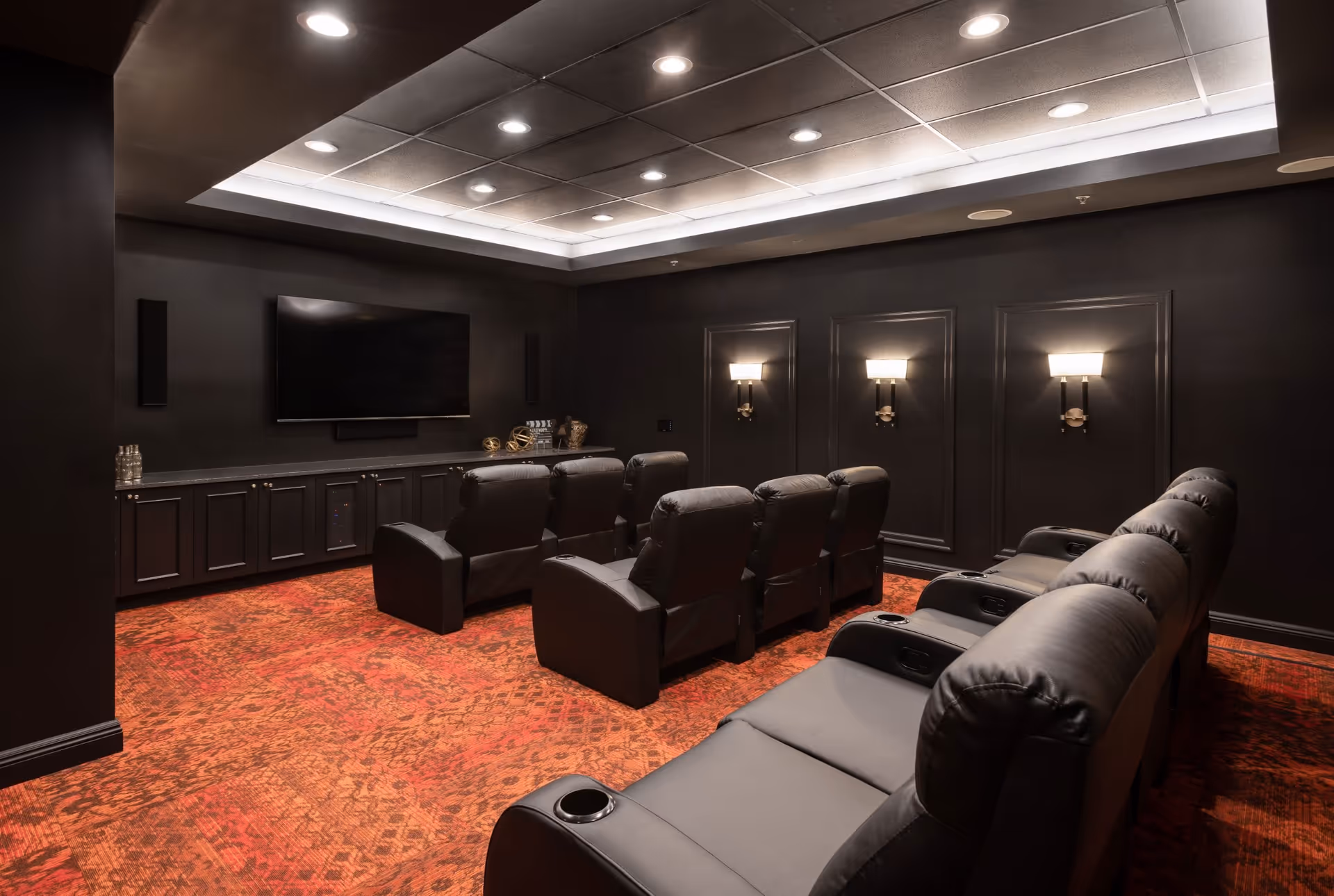 A home theater room with black leather recliner chairs arranged in two rows facing a large flat-screen TV mounted on a black wall. The room has dark walls, recessed ceiling lights, wall sconces, and a red patterned carpet.