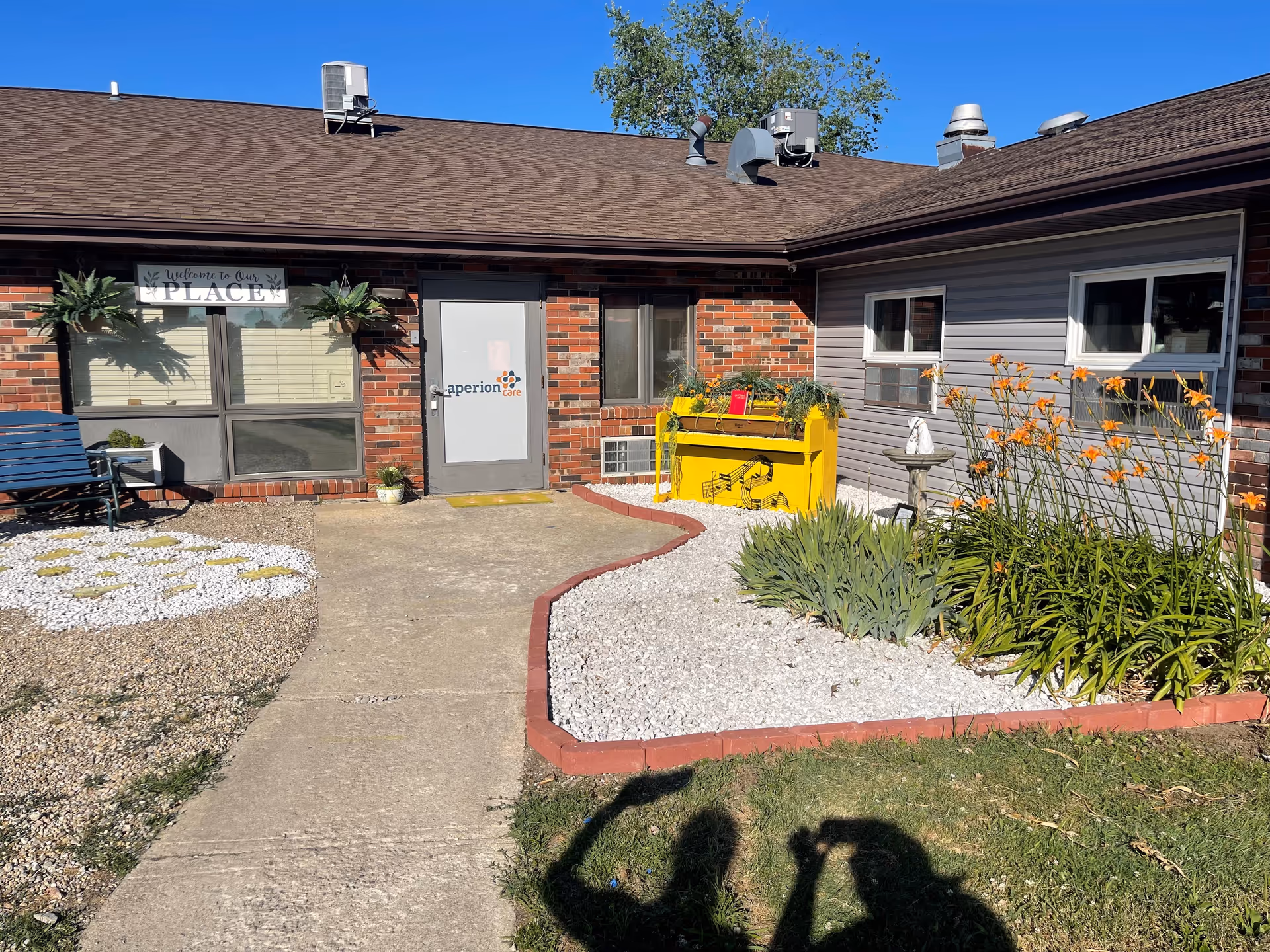 Entrance and front courtyard of a single-story brick and siding senior living facility with a paved walkway, a yellow planter, and orange flowers.