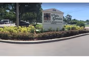 Outdoor view of the entrance sign for Palm Garden Health & Rehabilitation Center, surrounded by landscaped bushes and trees with a clear sky in the background.